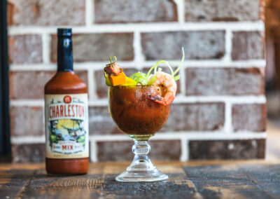 A bottle of Charleston Blood Mary Mix out of focus in the background. In the foreground is a goblet sized bloody mary stacked full of decorative food including a shrimp.