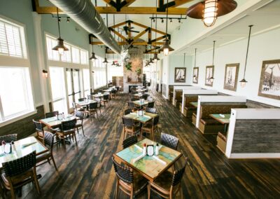 Beautiful and dramatic Fish House Indoor Dining Room with high ceilings decorated with structural beams and vintage style ventilation and craftsman lighting. A fireplace at the end of the great hall is flanked by booths and tables. Tables have nautical map inlays and wood. Images of large catch game fish in black and white are framed on the right far wall and across from that is a wall full of windows overlooking Charleston Harbor.