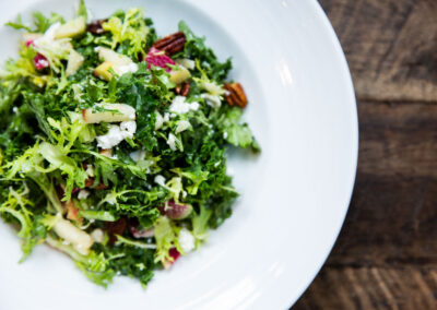 Mixed greens salad with walnuts tossed in and cheese crumbles. Resting on a round white plate that is centered on a rustic finished wooden table.